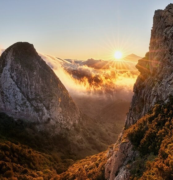 Los Roques de La Gomera al amanecer, con formaciones volcánicas y un valle cubierto de nubes y vegetación.